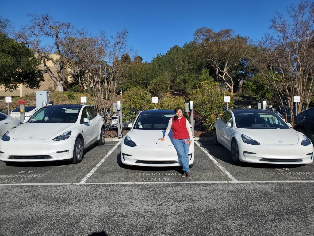 A woman wearing a red vest and white shirt posing in front of three white Teslas.