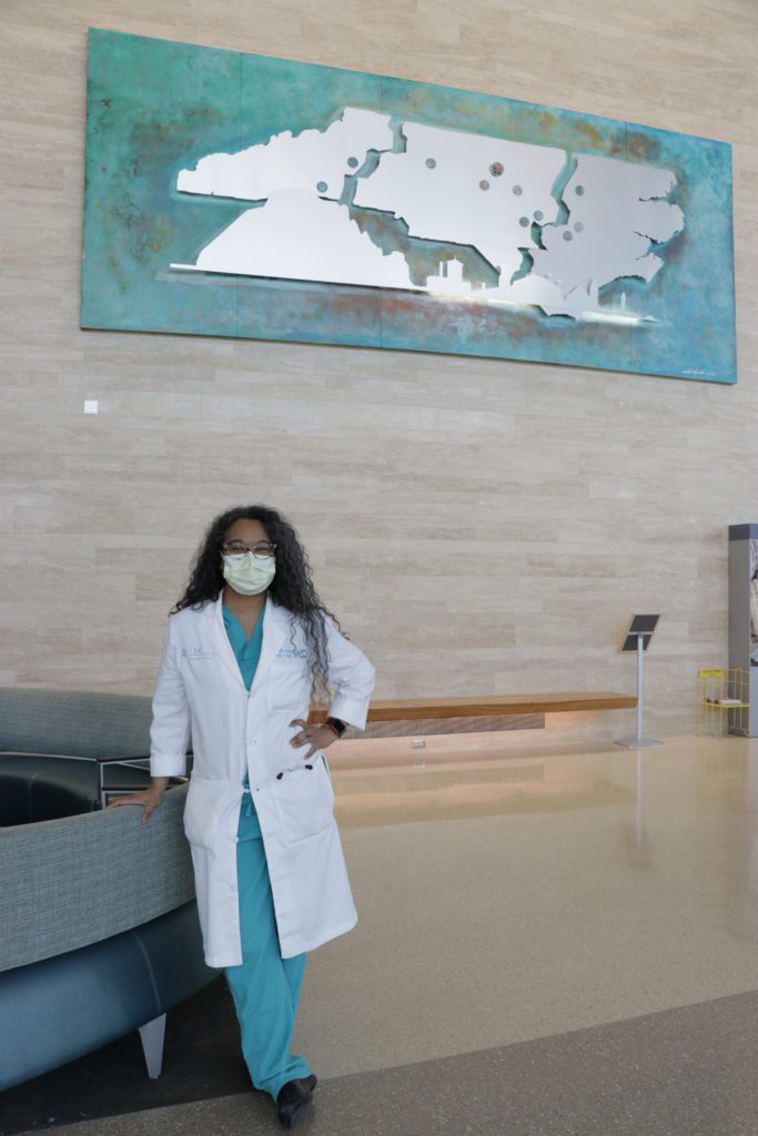Woman wearing a white lab coat and scrubs standing in front of a sign at the University of North Carolina Chapel Hill Department of Surgery