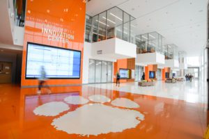 The entrance of the Watt Family Innovation Center displays the Clemson Tiger Paw on the floor and a spacious lobby with five large digital screen that lead from entrance to exit. Many SAS software projects are housed here.