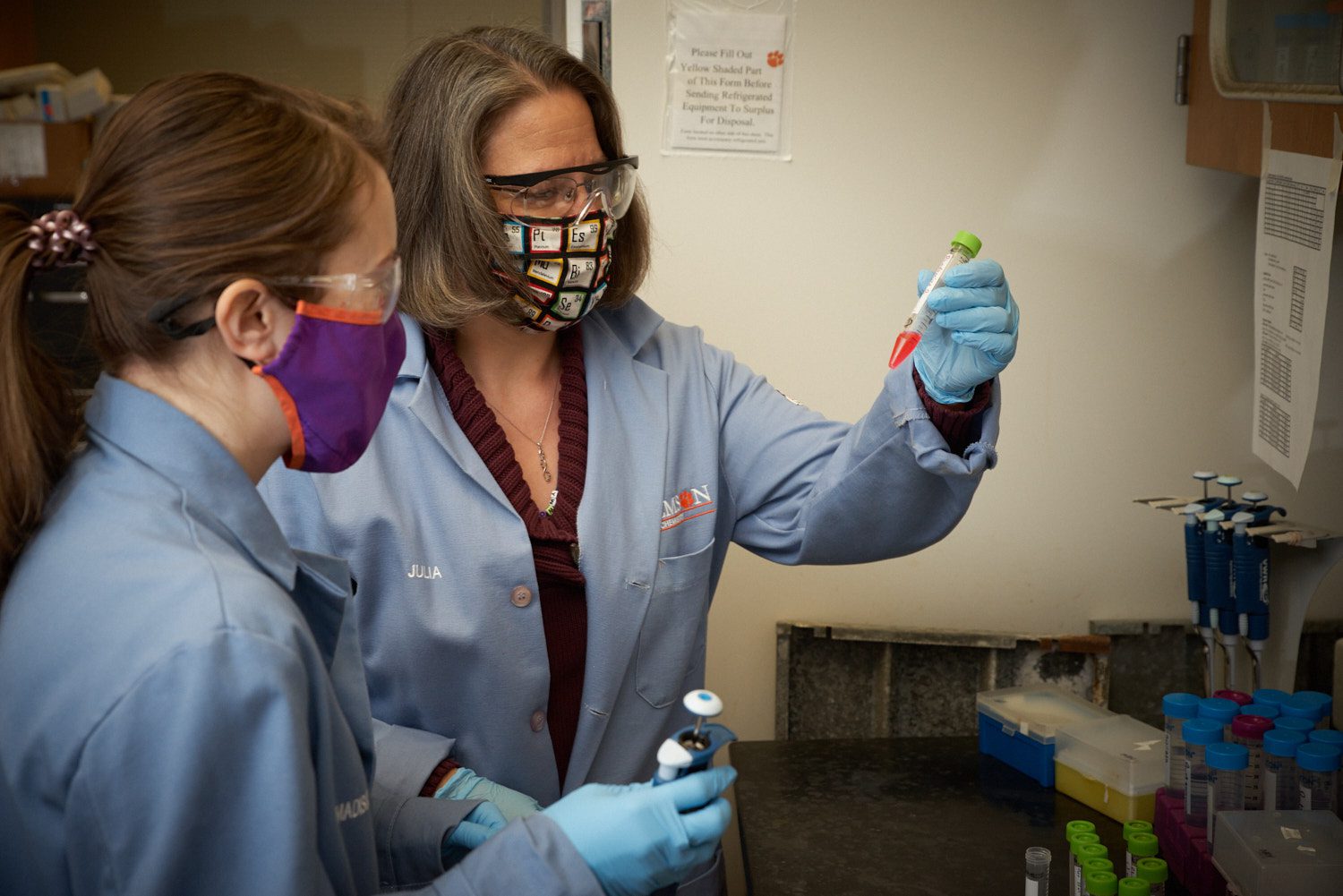 Woman wearing a blue lab coat and a periodic table mask holds a test tube with a red substance in it as another woman in a blue lab coat looks at it.