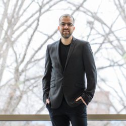 A smiling latinx man standing in front of a glass wall wearing a suit with hi-top sneakers. His hands are in his pockets. His is an economic assistant professor at Clemson University.