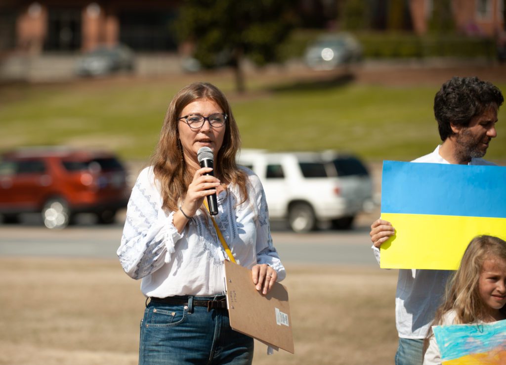 Lyudmyla Tsykalova, a recent Ph.D. graduate of Clemson’s International Family and Community Studies program, speaks into a microphone from Bowman Field, with people holding Ukrainian flags in the background.