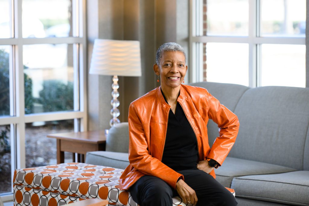 A smiling African American woman with short hair sits on the end of an ottoman with a sofa, end table and lamp in the background. The room has floor to ceiling windows behind the sofa and also on the wall parallel to the side table and ottoman. She is wearing dark jeans and a sweater with a bright leather jacket. Her dangling teardrop earrings match her leather jacket. She received the Distinguished Service Award. 