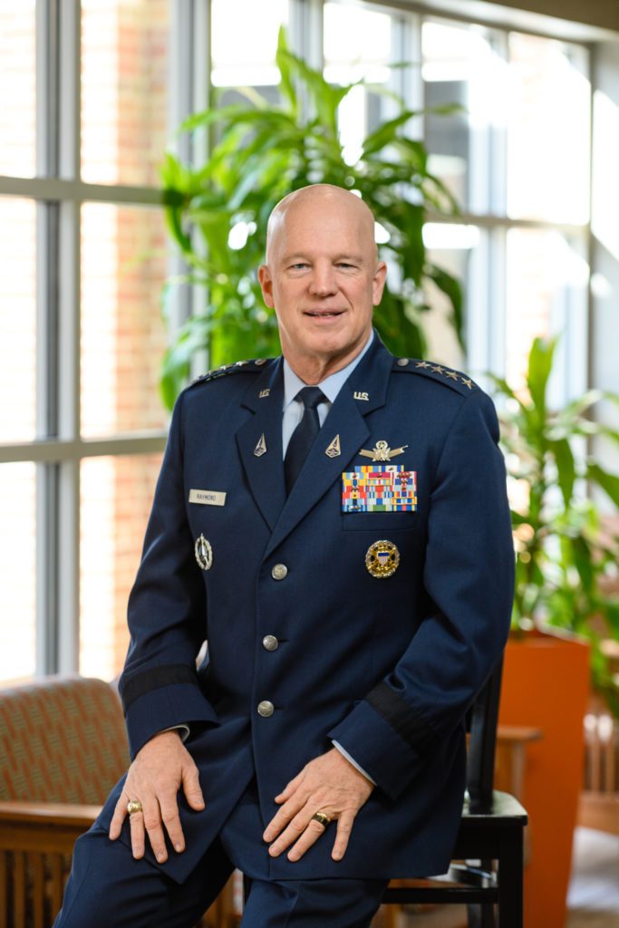 In a highly decorated Air Force service dress uniform, a man sits/leans against the arm of a chair. Both of his hands are placed on each corresponding leg. He has a smile and no hair. He is wearing a wedding ring and his Clemson class ring. He received a Distinguished Service Award.