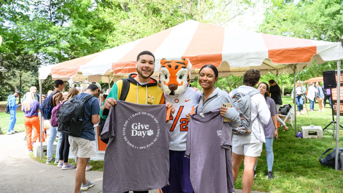 A multi-racial male student and female student pose with the Tiger Cub mascot while holding Give Day t-shirts they received from making a gift. Behind them, on the lawn, stands a tent over a table and smiling students all around.