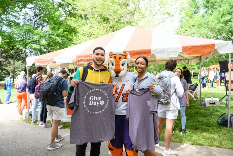 A multi-racial male student and female student pose with the Tiger Cub mascot while holding Give Day t-shirts they received from making a gift. Behind them, on the lawn, stands a tent over a table and smiling students all around.