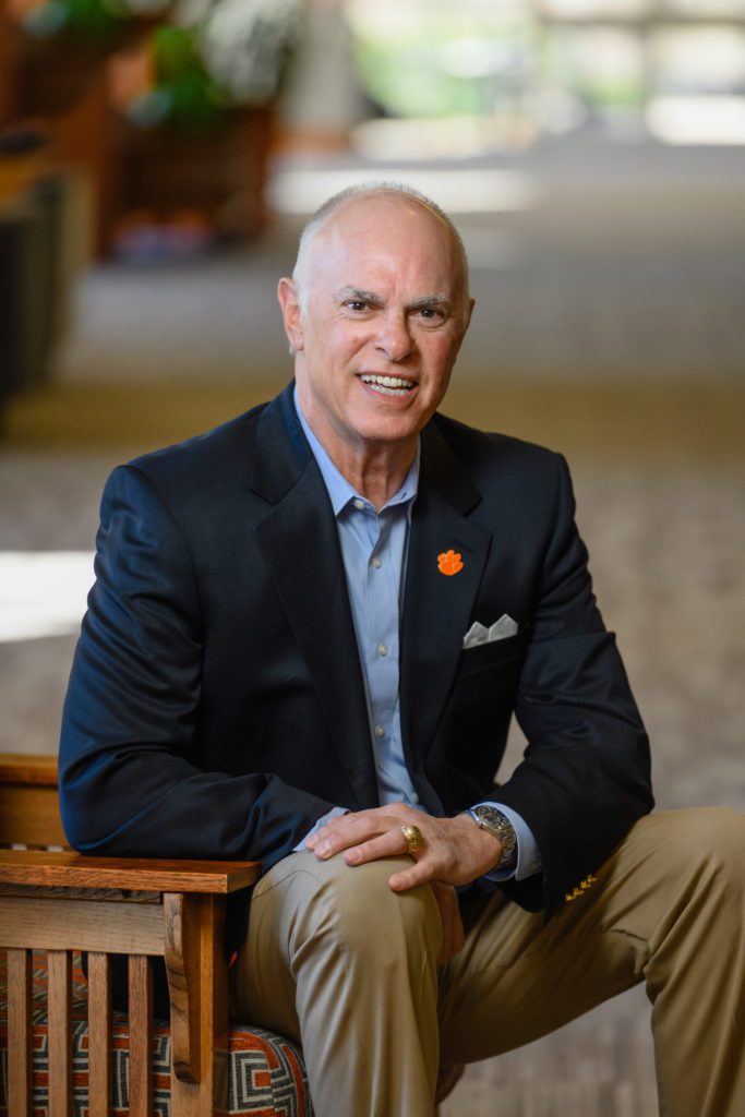 A smiling man sitting on the edge of a chair the background is a hallway. He isn't completely bald. He is wearing dress slacks and a blazer with a Clemson Tiger paw pendant. He received a Distinguished Service Award.