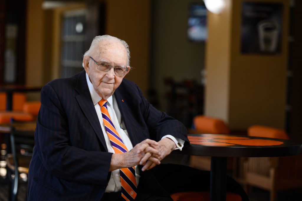 An older man sits at a table that features the Clemson Tiger Paw logo on the surface top. He is wearing a suit and glasses, hands clutched within one another. His hair is white from years and barely visible on the top and sides. He has a slight and a very distinguished demeanor. He was a warded a Distinguished Service Award.