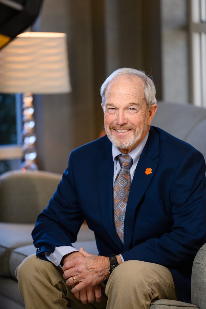 A smiling man with a mustache and short beard, hair thicker on the sides than top, sits comfortably on a sofa with a table and lamp behind him. He is wearing khakis,  a blazer and tie with a Clemson Tiger Paw pendant. He received a Distinguished Service Award. 