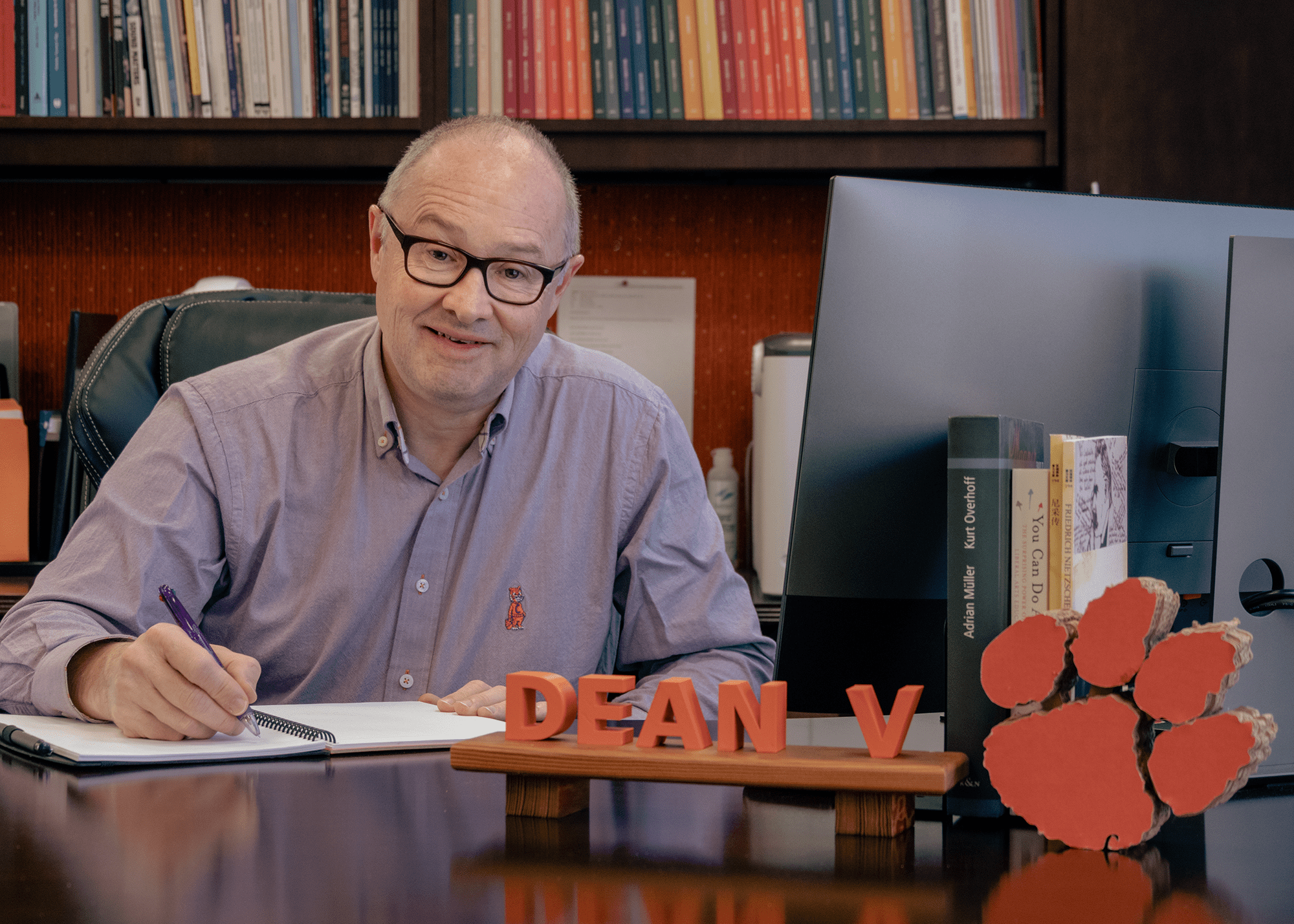 Nicholas Vazsonyi at desk, smiling, with pen in hand. Sign on desk says "Dean V." Large orange Clemson Tiger paw is on his desk.