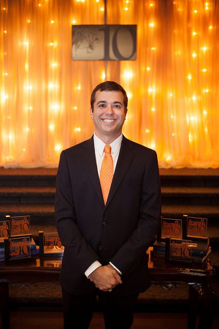 A smiling man of Caucasian descent in a suit with a neck tie. His hands are clutched in front of his suit. In the background is a stage with light-weight curtains that are lined with string lights from the floor to the ceiling. He is a 2022 Clemson Alumni Association Board Member.