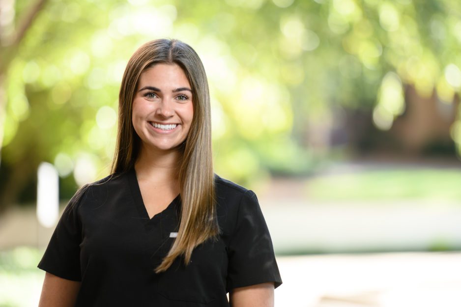 A brunette female student in black scrubs poses outside on campus.