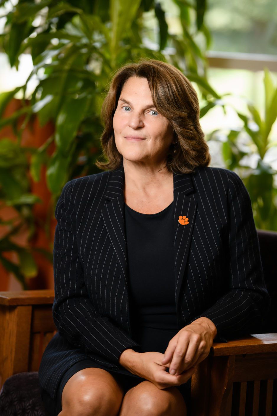 A smiling woman in a dress suit, sitting in a wood chair, one arm leaning on the arm of her chair and the other in her lap, behind her are plants. She, dean Hossfeld, is an expert in food insecurity.