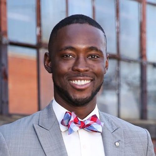 A smiling man of Black descent with short hair and a low-cut beard and moustache, wearing a suit with a bowtie. He is standing outside in front of a wall of tall, industrial windows. He is a 2022 Clemson Alumni Association Board Member.