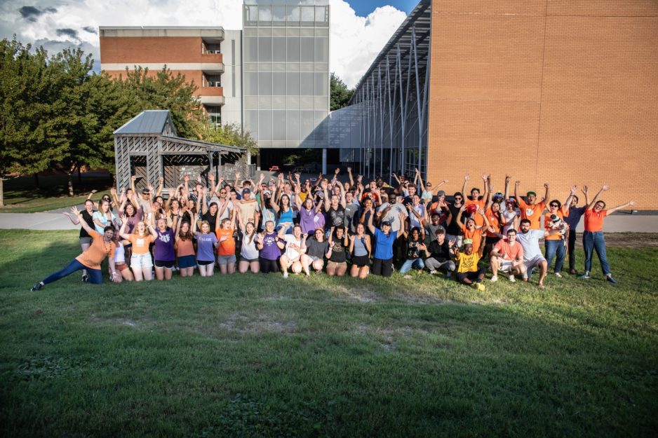 dozens of architecture students wearing bright, casual clothing raise their hands in celebration for a group photo. They sit in a green lawn in front of a modern brick and glass building at sunset.