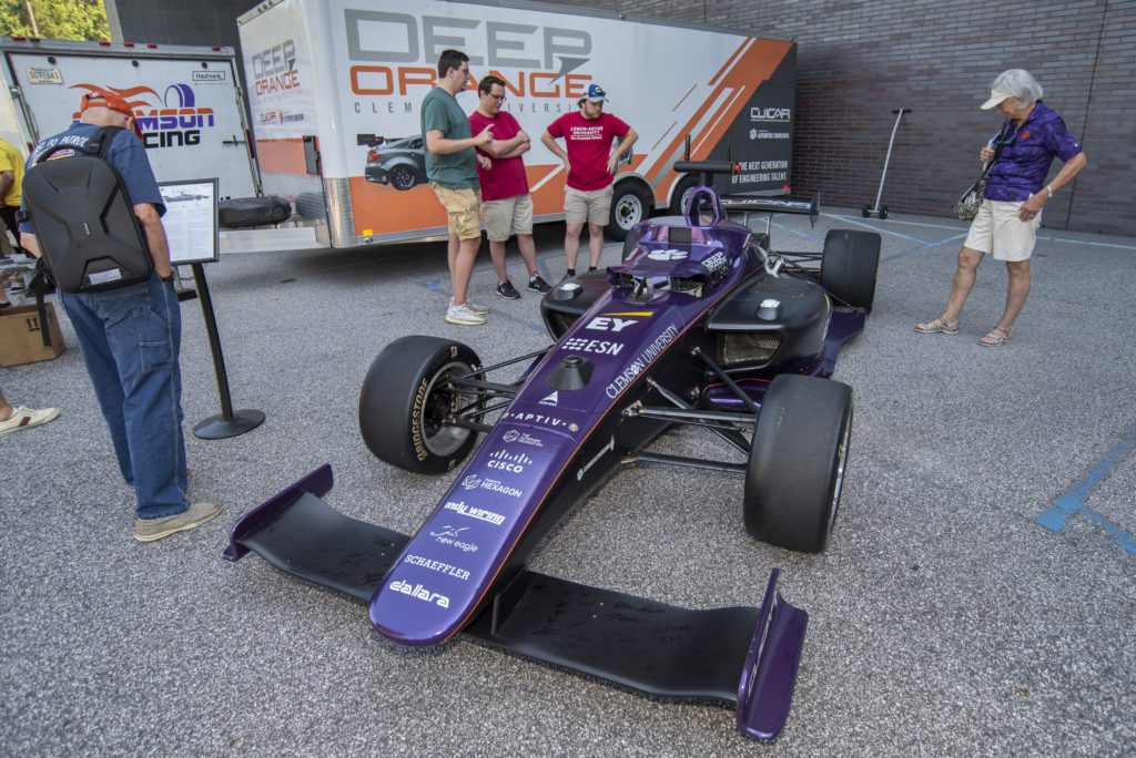 Three people look at the purple and black Indy car on display.