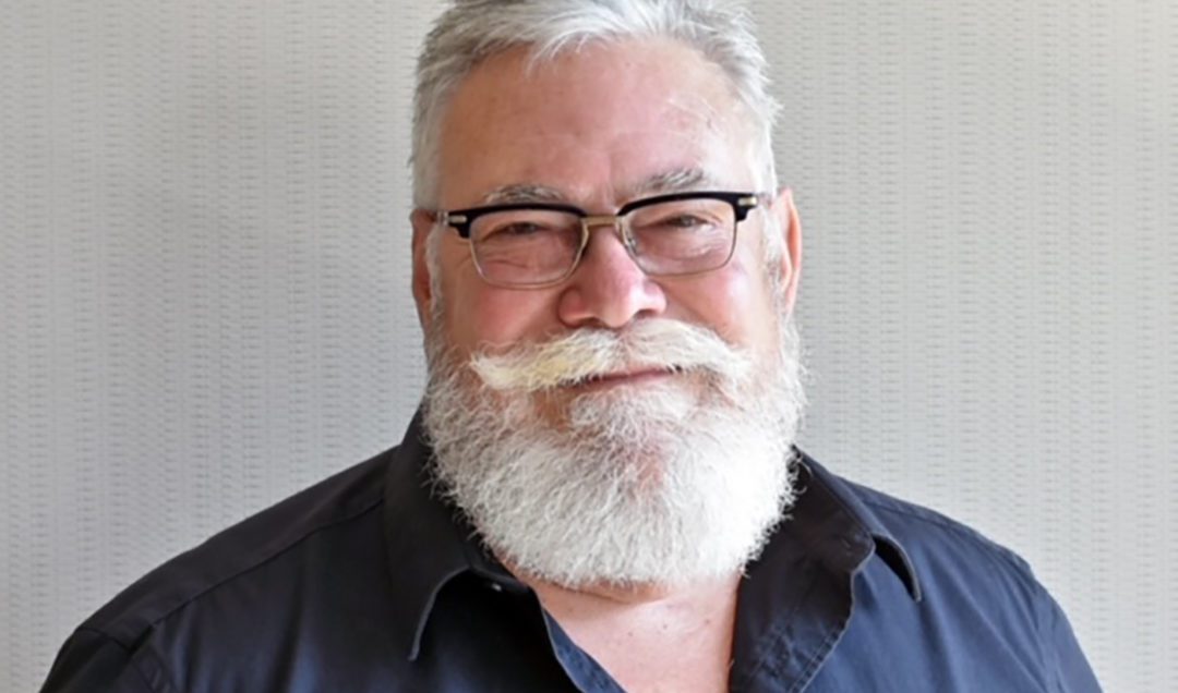 Photo of Mark Hayward, a man with white hair and beard, dark shirt and glasses in front of a white wall.