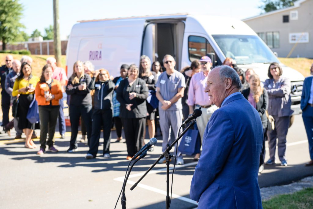 Sen. Thomas Alexander speaks into a microphone at a podium while about 20 people look on with a Clemson mobile health unit in the background.