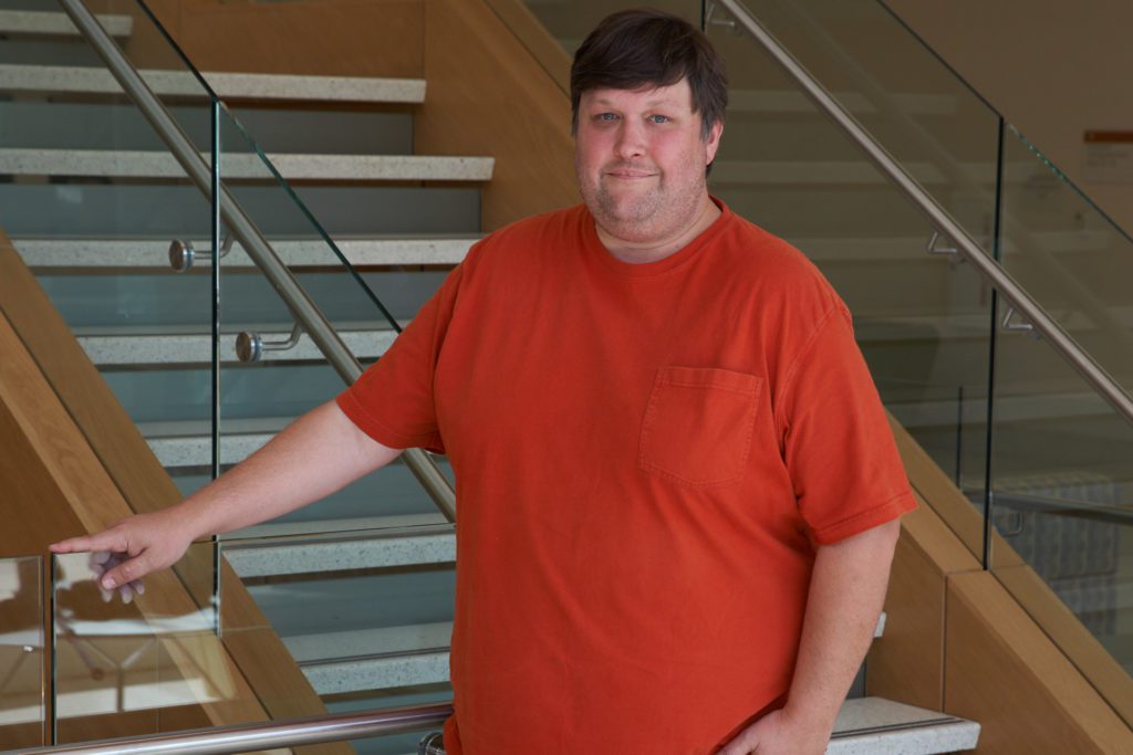 Man wearing orange shirt (Dan Whitehead) standing near stairs in Life Sciences Facility