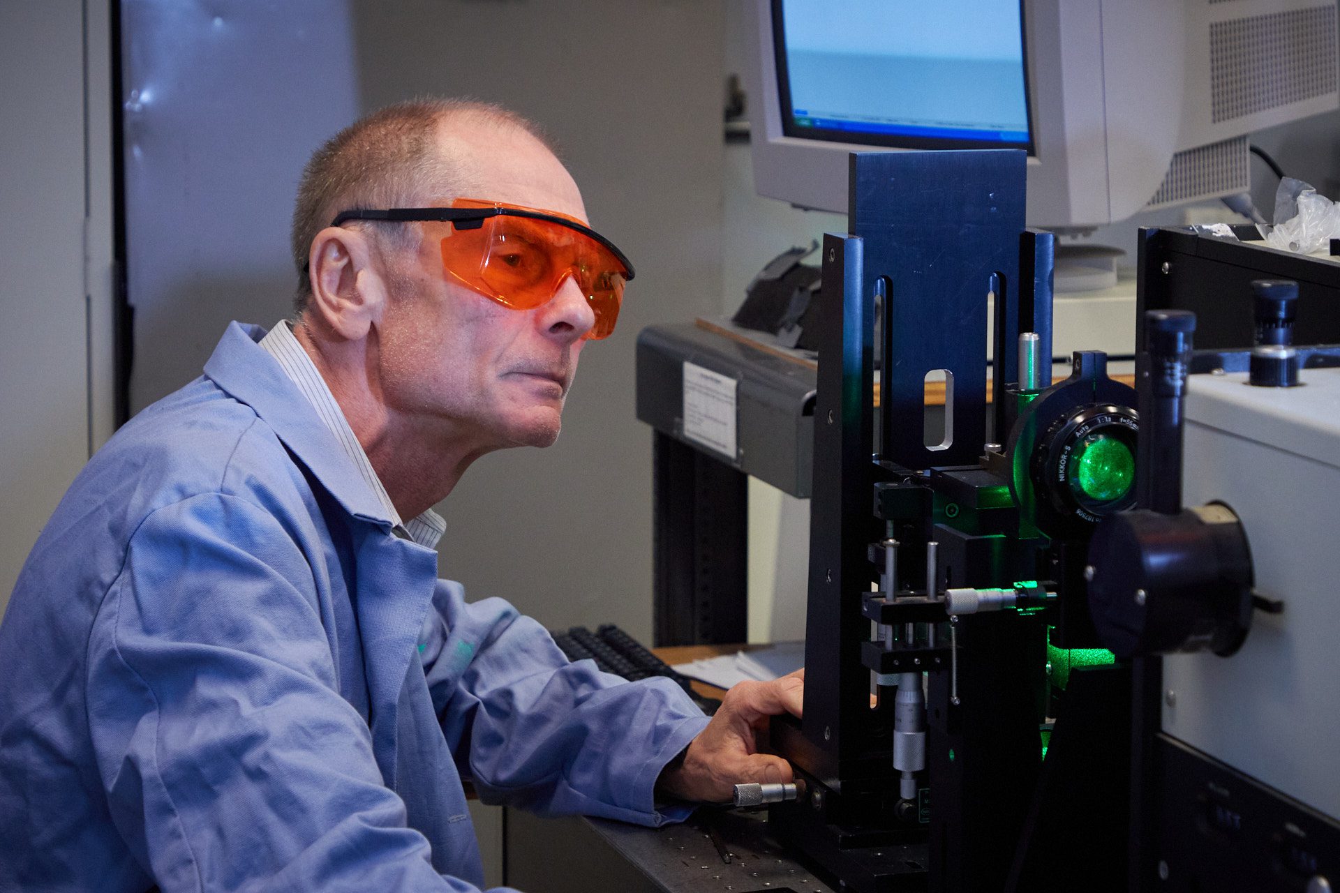 Man wearing safety goggles and blue lab coat looks at machine in lab.