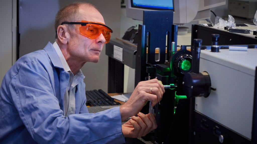 Man wearing goggles and a blue lab coat works with a machine with a green light in a lab