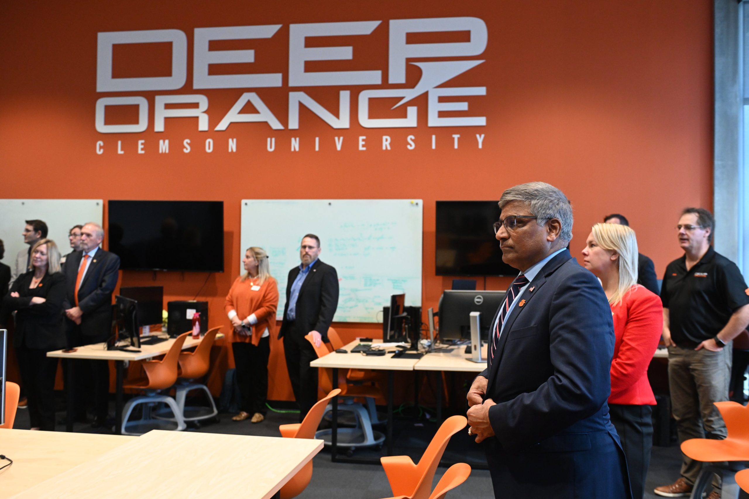 Ten people, including the NSF Director, are standing by a number of tables at the Clemson ICAR campus. Inside the multi-stories room is a background wall with the words "Deep Orange" printed on the wall.