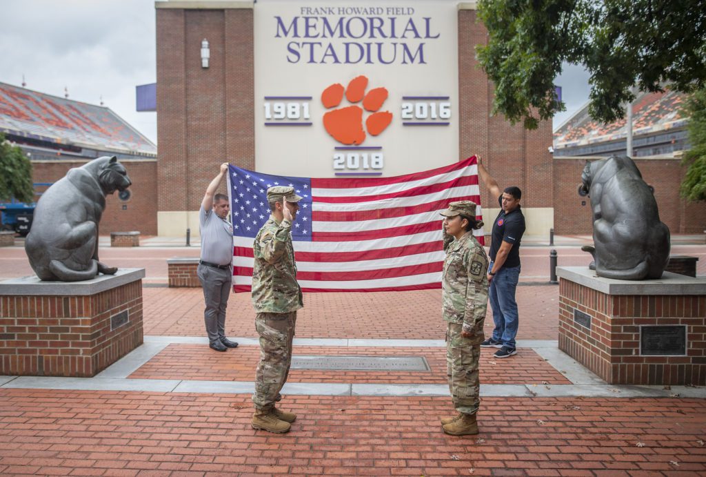 A woman and man wearing U.S. Army uniforms raise their hands in a salute to each other. Behind them are two men carrying a U.S. flag and the signage of Memorial Stadium. 