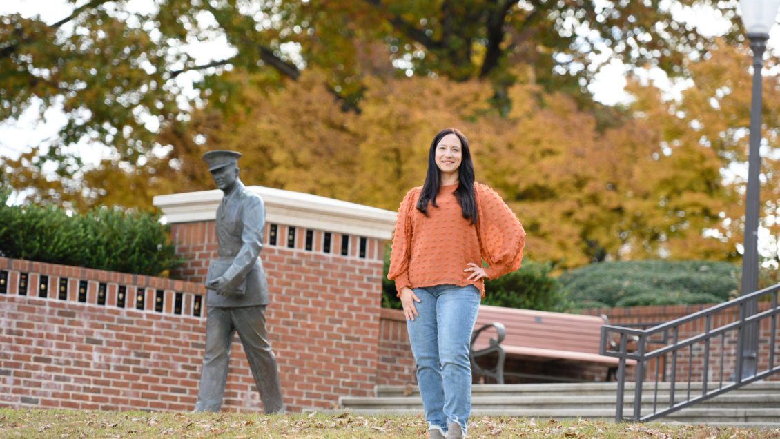 Biological sciences student Rachelle Hales wears an orange sweater and jeans and is standing with her hands on her hips in a plaza with a military statue.