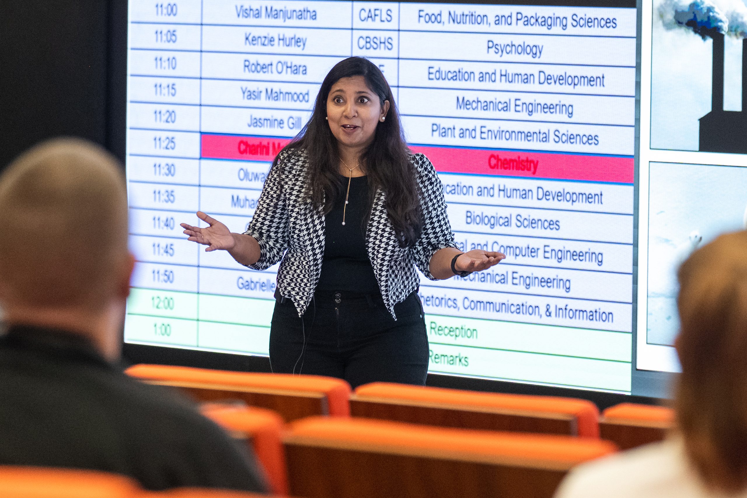 A woman presents in front of a large screen displaying a table with various categories listed. Chemistry is highlighted in red.