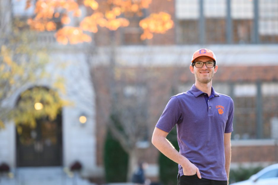 A male student wearing an orange baseball cap and purple polo shirt poses outside of Riggs Hall.