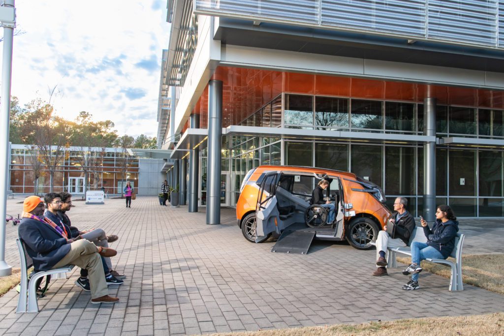 An orange pod-shaped car has its doors open, revealing its wheelchair-accessible capabilities.  Five people sit on two benches near the car. 