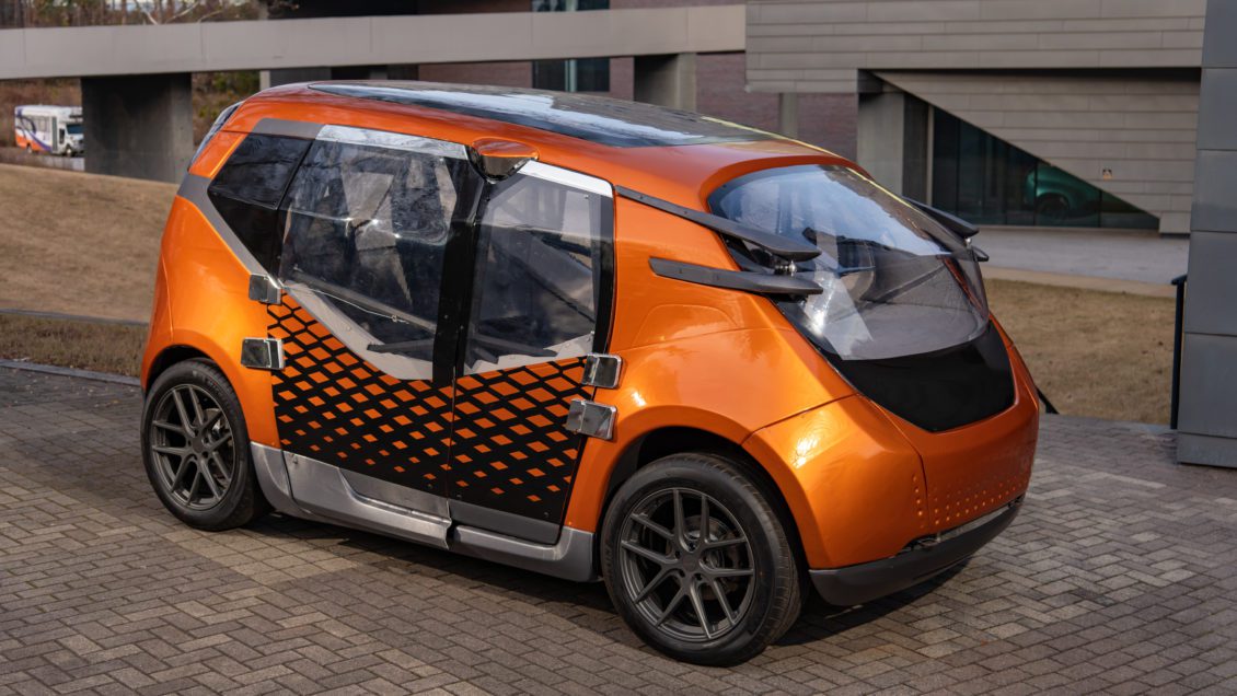 A futuristic orange car sits in an empty brick parking lot of a commercial complex.