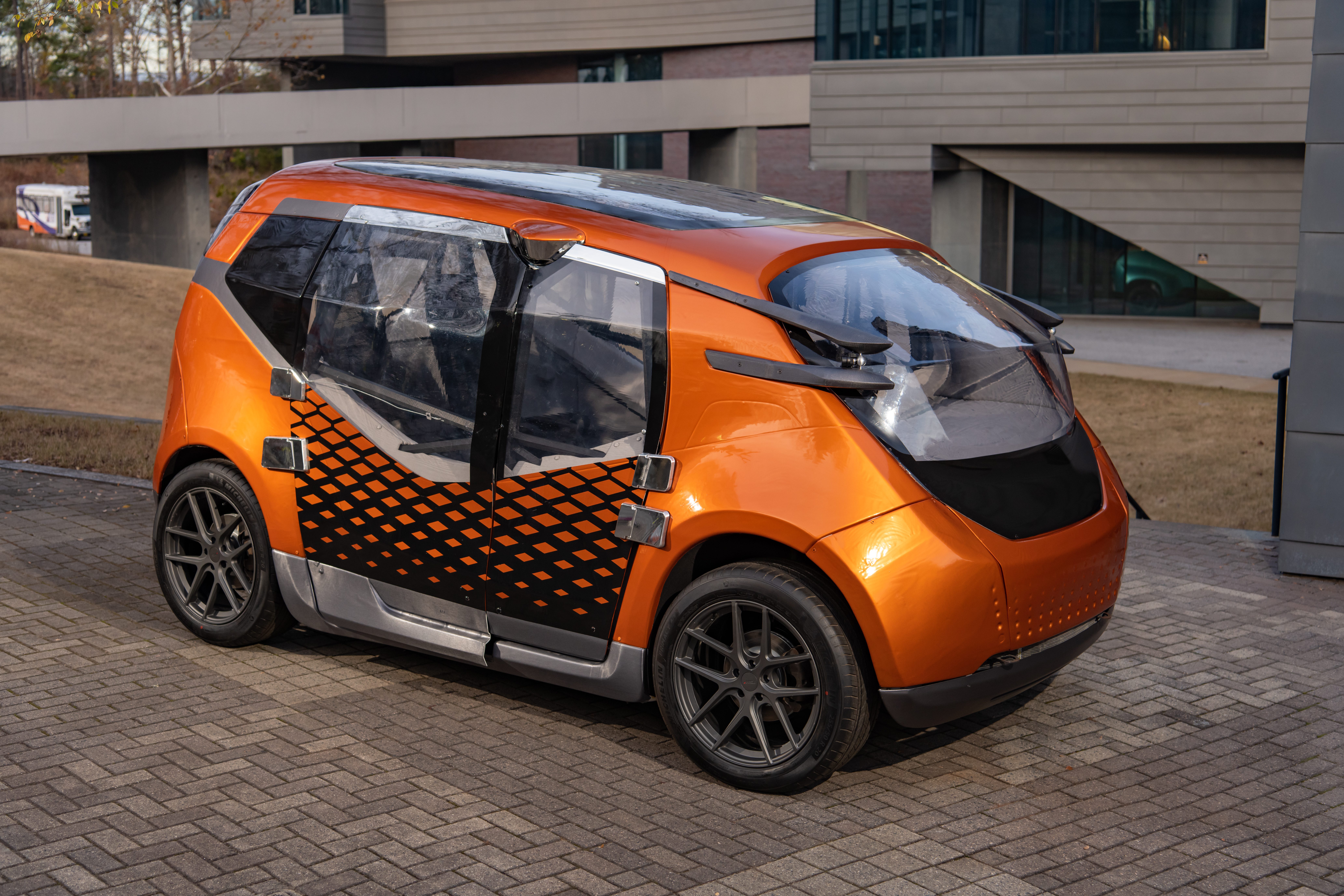 A futuristic orange car sits in an empty brick parking lot of a commercial complex.