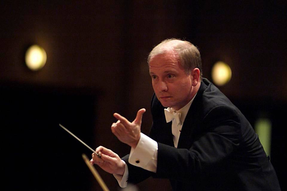 A conductor wearing a formal black suit waves his baton and gestures 