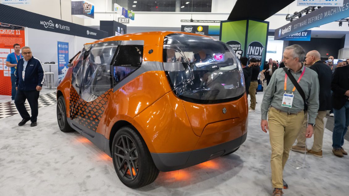 The Deep Orange 11, an orange vehicle, sits in a large space surrounded by people wearing lanyards as they view the car.