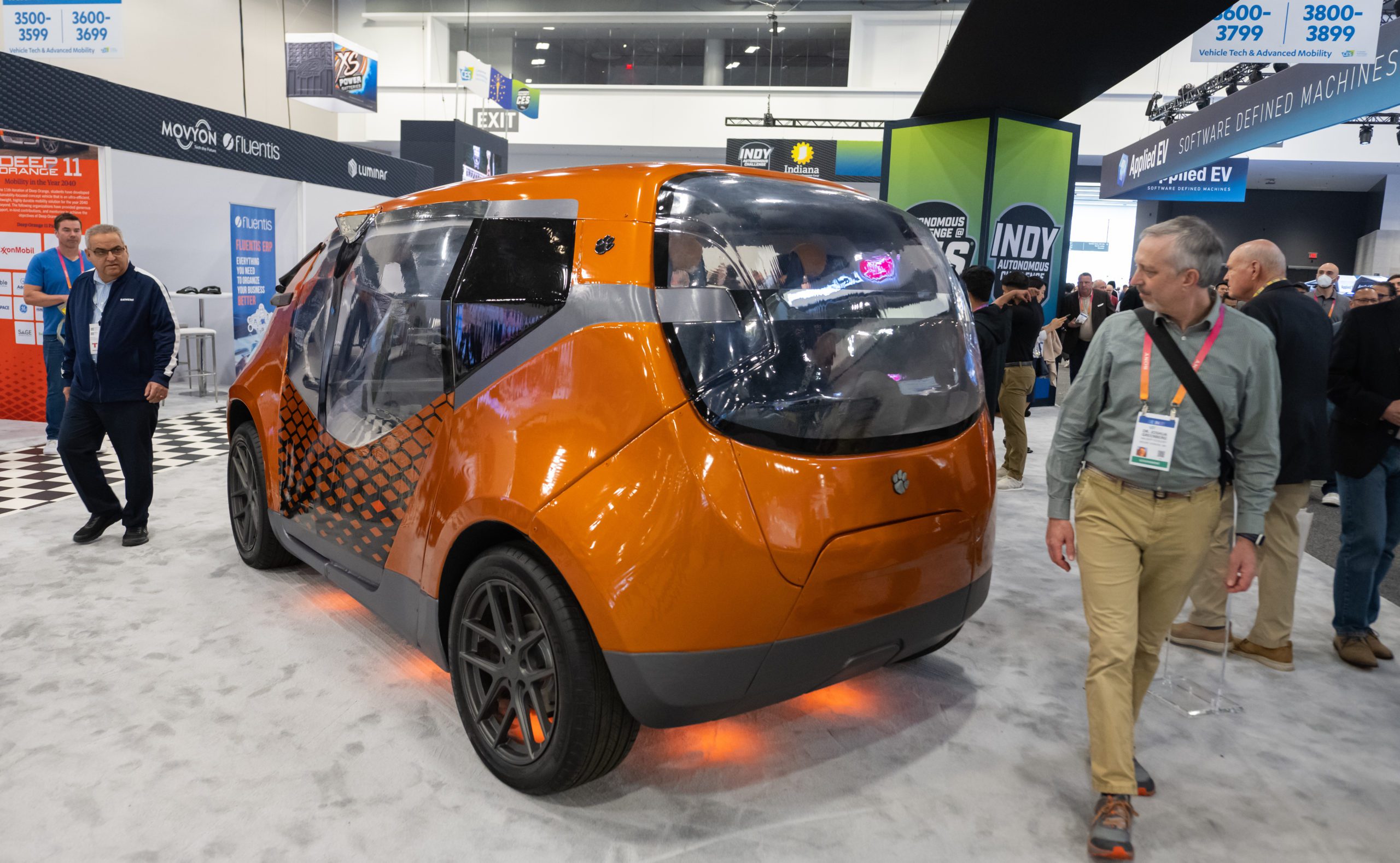 The Deep Orange 11, an orange vehicle, sits in a large space surrounded by people wearing lanyards as they view the car.
