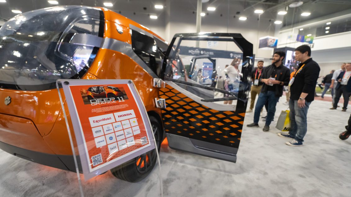 An orange futuristic concept car with an open door is surrounded on the floor of the Consumer Electronics Show floor has several people looking at it.