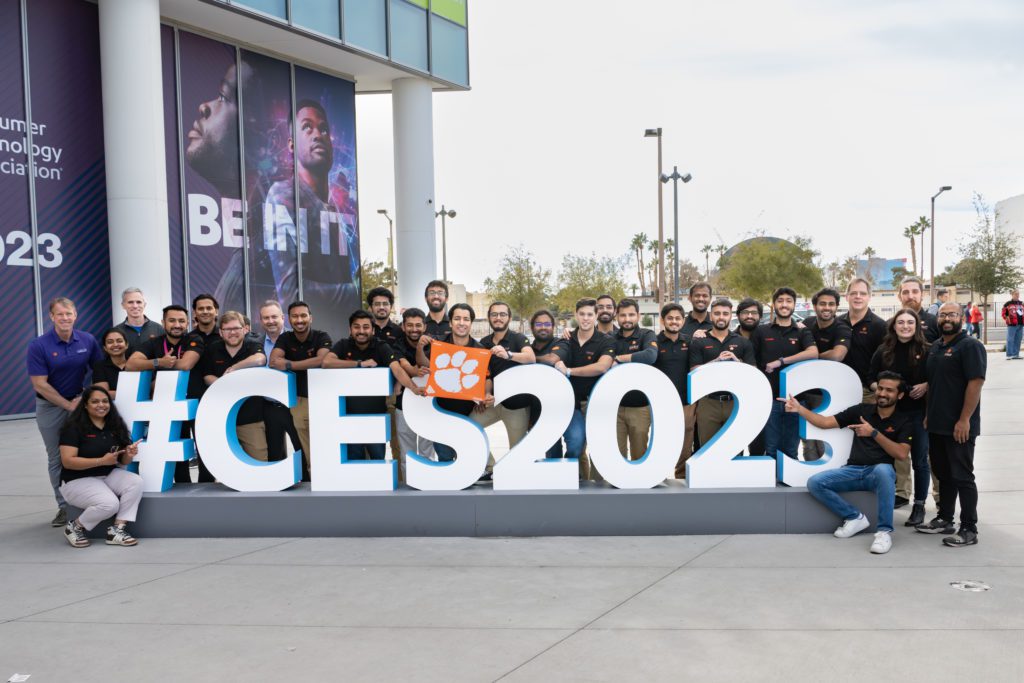 A group of students wearing black, branded Clemson shirts hold a Clemson Tiger Rag while gathered around a huge CES2023 sign outside a large commercial building.