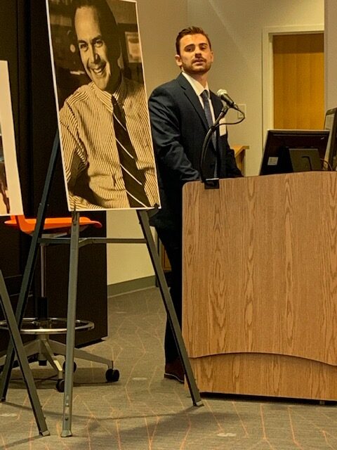 A younger caucasian man, former student, dressed in a suit, is standing behind a podium speaking, and looking over at the poster-sized photo of the man that had been his mentor, Ben Dysart.