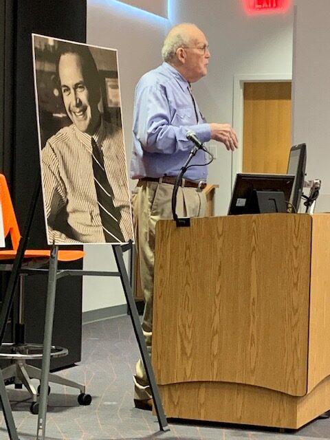 Older caucasian man standing behind a podium, speaking. To his right is a poster-sized photo of him, Ben Dysart, when he was a much younger man.