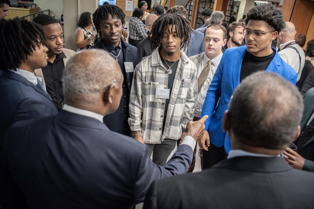 A man with his back to the camera speaks to a group of young men, pointing at one of them