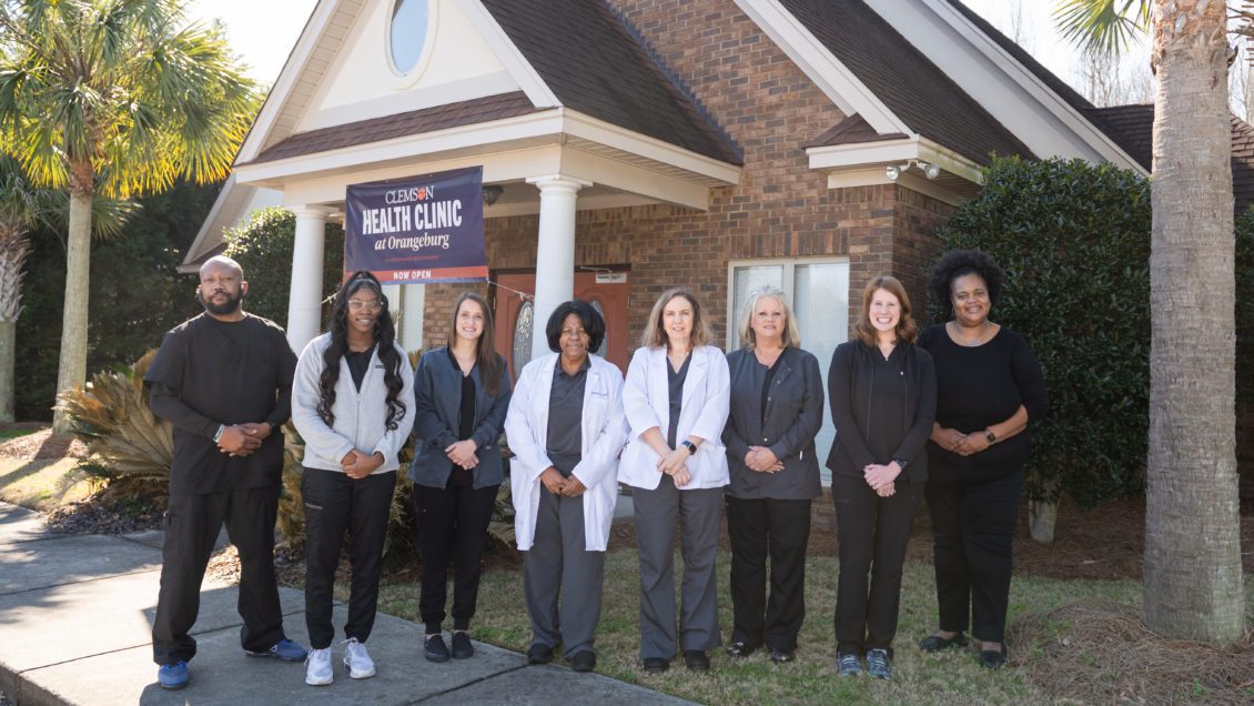 A group of health care providers wearing scrubs and white coats stands outside a brick medical facility.