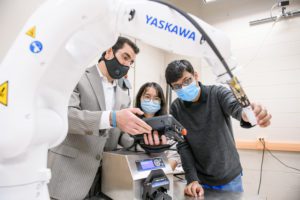 Three individuals wearing masks control a robotic arm with a hand-held device. 