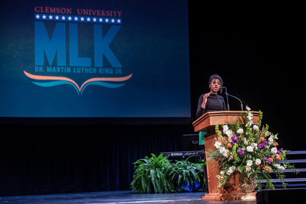 A young woman speaks from a podium in front of a projected graphic of "MLK"
