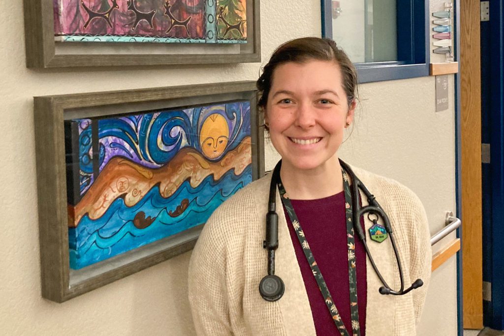 A woman (Katie Gray) with a white lab coat and stethoscope standing in front of some Alaskan native art.
