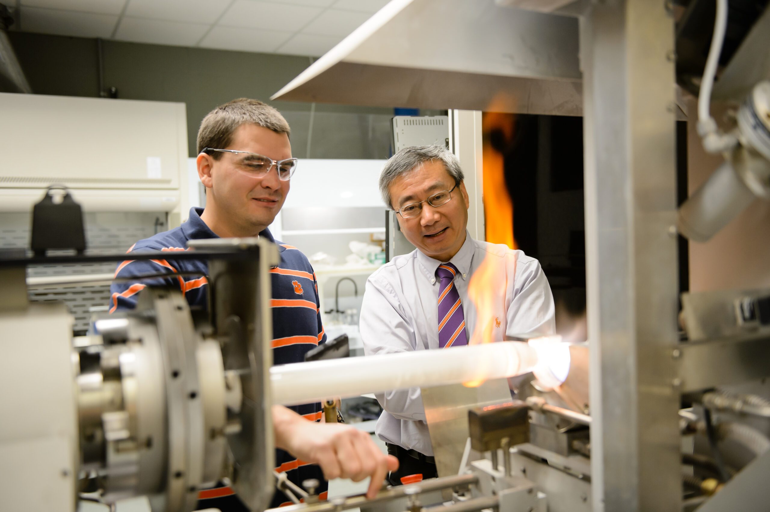 An Asian Clemson professor wearing glasses and a purple and orange tie stands next to a male student wearing safety goggles and a Clemson polo shirt, and a tube of light with flames coming off of it is in the foreground. 