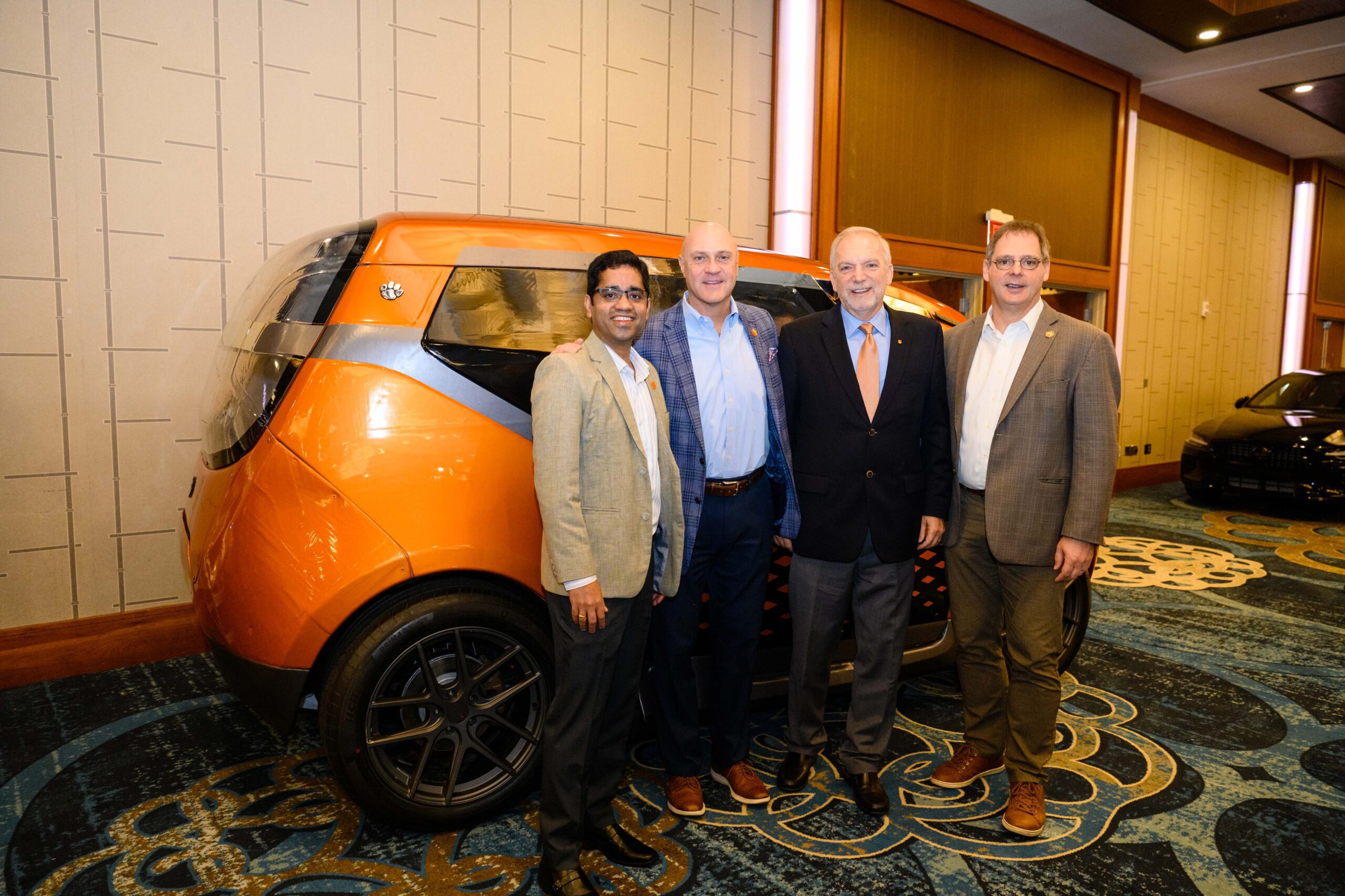 Four men wearing coats and collared shirts stand in front of an orange, futuristic car located inside an indoor, carpeted space, the S.C. Automotive Summit.