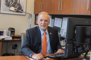 A man wearing a dark blazer with a Clemson Tiger Paw lapel pin and an orange tie sits behind a desk with a computer in front of him. 