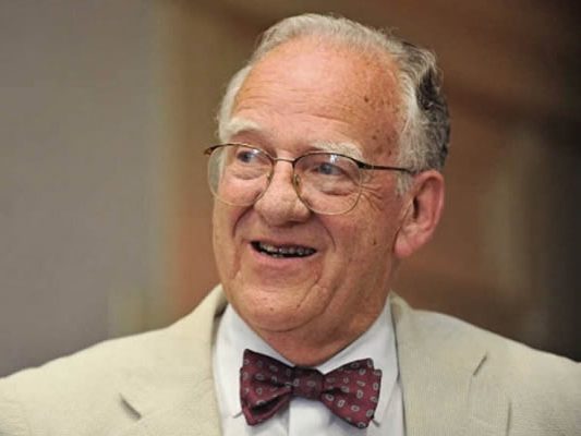 A head and shoulders shot of an older white man, Bruce Yandle, is looking to the side and has a smile on his face. He is wearing a bow tie, blazer and glasses.