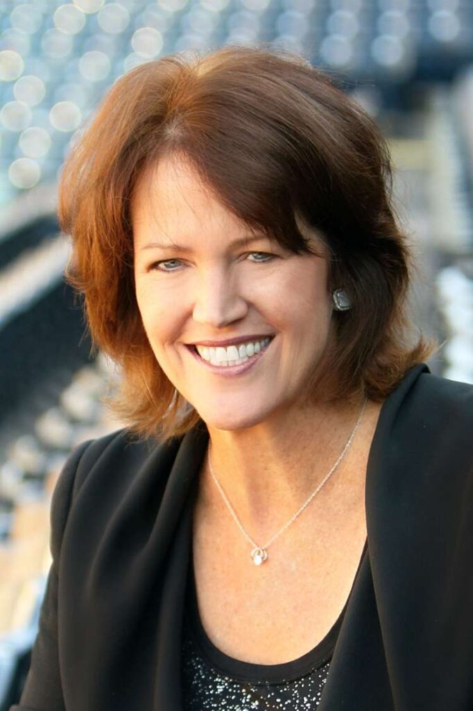 Christine Brennan, a woman with shoulder-length hair parted in the middle with slight bangs is smiling. Shown from the shoulders up, she is wearing a blazer and shell. A necklace with a small emblem dangles from her neck. The background is a sports stadium. 